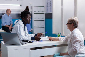 African american doctor consulting old woman at desk in healthcare cabinet. Black medic discussing health problems with sick elder patient at clinic with medical equipment and tools