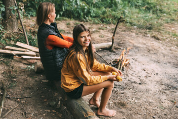 A mother with her teenage daughter is sitting on a tree near a campfire in the forest or park during a picnic. Weekends, vacations, time together