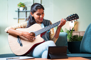 Young girl learning to play acoustic guitar at home from digital tablet through video call - concept of new normal, online or virtual education during coronvirus covid-19 pandemic.