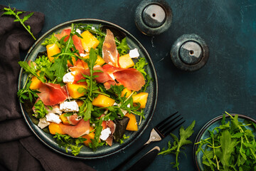 Fresh salad with cantaloupe melon, prosciutto, soft cheese and arugula on blue background, top...
