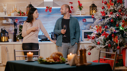 Couple clinking glasses of champagne chatting after christmas dinner. Man and woman standing in festive decorated kitchen drinking alcohol and enjoying holiday celebration. Winter festivity