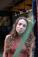 European girl with dark and curly hair with restaurant behind and palm leaf in front