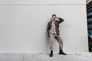 Fashionable young man in stylish casual look clothes with oversized blazer, shirt and black shoes stands near a gray wall in the city - Powered by Adobe
