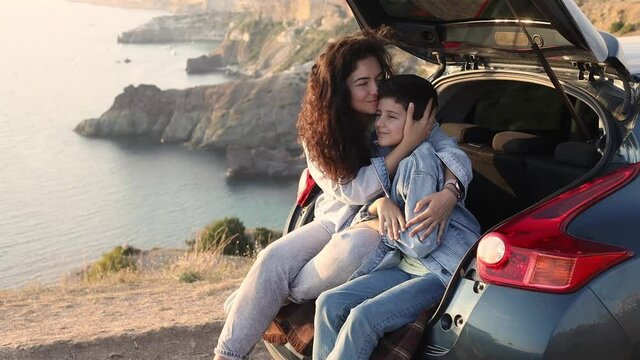 A young mother and her teenage son in denim enjoy the view of the sea and mountains on the top of the hill, sitting in the open trunk of a car and hugging together. Happy family relationship concept. 
