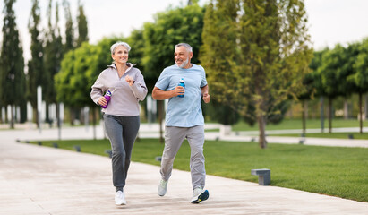 Full-length photo of lovely joyful retirees couple jogging outside in city park