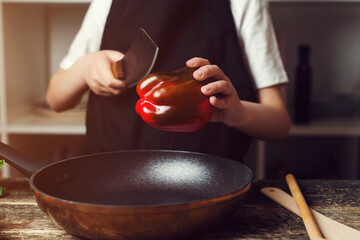 Boy cutting red pepper for cooking dinner. Chef wearing apron at the kitchen. Cooking food concept.