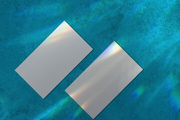 Two empty white rectangle a4 poster, business card or flyer mockups lying diagonally with overlay of rainbow light refraction caustic effect and shadow on trendy mint blue concrete background.