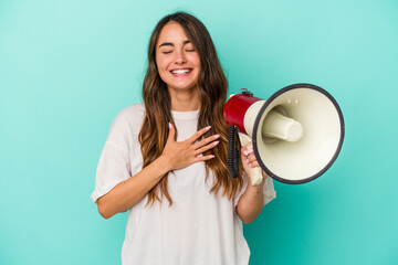 Young caucasian woman holding a megaphone isolated on blue background laughs out loudly keeping hand on chest.