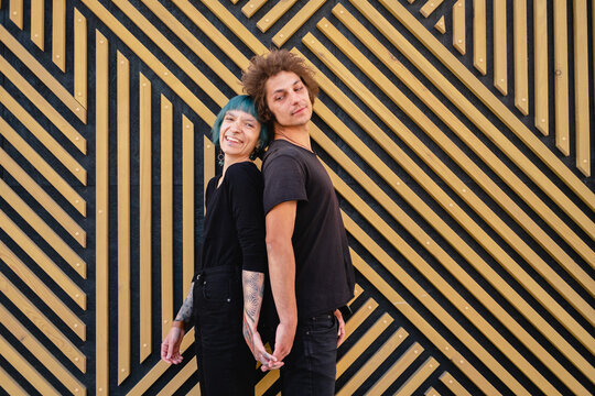 Happy emotional couple of young man and woman in black casual clothes standing back to back on a wooden wall background.