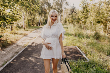 Happy pregnant woman outdoors on a sunny day, smiling, wearing a white mini dress.