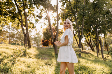 Pregnant woman walking in a park on a sunny day, smiling, wearing a white dress.