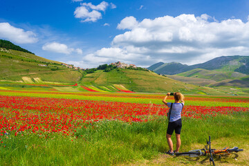 Fototapeta na wymiar Woman cycling mtb in blooming cultivated fields of Castelluccio di Norcia highlands, famous colourful flowering plain in the Apennines, Italy. Agriculture of entil crops and red poppies.