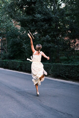 Young woman in dress and hat running and jumping down a road surrounded by trees