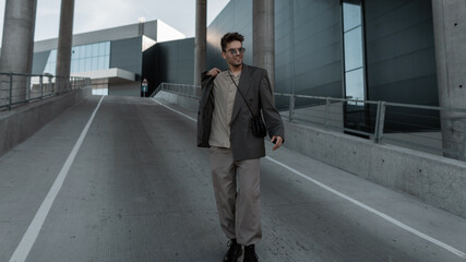 Happy handsome hipster man with sunglasses in fashion gray suit with handbag walking along the road in the city