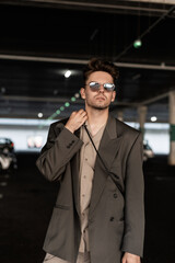 Stylish male businessman model with sunglasses in fashion blazer and shirt walk on parking lot