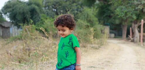 A cute little Indian boy standing in the field smiling looking at the ground in painted green t-shirt