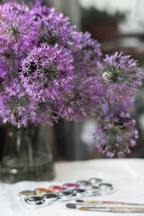 bouquet of blooming decorative onions of violet-lilac color in a glass vase stands on the table next to brushes and watercolor paints