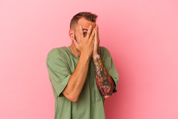 Young caucasian man with tattoos isolated on pink background  blink through fingers frightened and nervous.