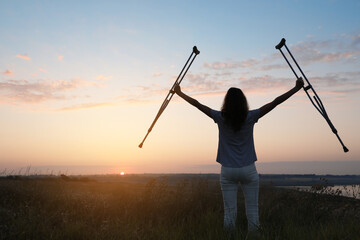 Woman holding axillary crutches outdoors at sunrise, back view. Healing miracle