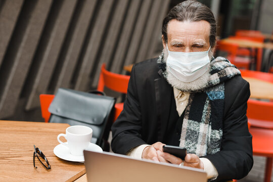 Senior man in face mask using cellphone while working with laptop