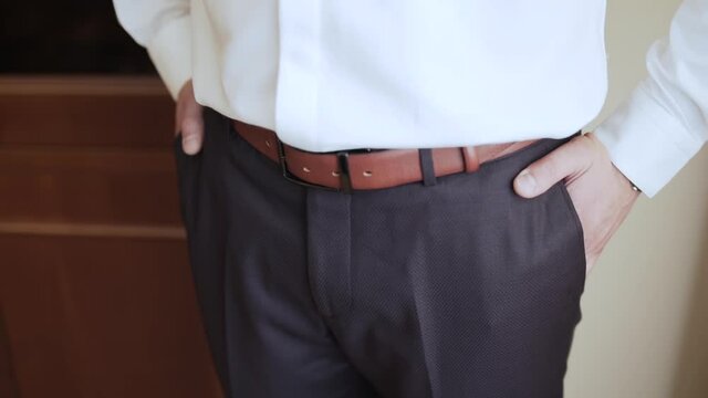 Man in white shirt and brown trousers with belt puts his hands in his pockets at home, close-up. Morning of a businessman.