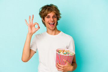 Young caucasian man with makeup holding popcorn isolated on blue background  cheerful and confident showing ok gesture.