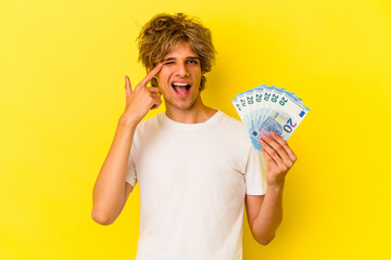 Young caucasian man with makeup holding bills isolated on yellow background  showing a disappointment gesture with forefinger.