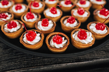 Close-up dough tartlets with cheese cream, cherries stand on a round tray, plate on a wooden table. Banquet, buffet, top view.