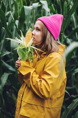 Funny little girl dressed in a yellow raincoat and a hot pink cap spoils and bites corn in a cornfield