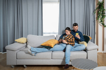 Obraz premium Young married couple in their home internet and social media networks addicts surf the internet on their phones and they are completely uninterested in each other