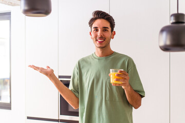 Young mixed race man drinking orange juice in his kitchen showing a copy space on a palm and...