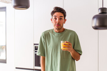 Young mixed race man drinking orange juice in his kitchen shrugs shoulders and open eyes confused.