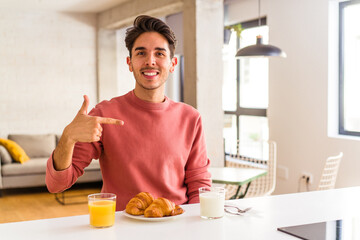 Young mixed race man having breakfast in a kitchen on the morning person pointing by hand to a shirt copy space, proud and confident