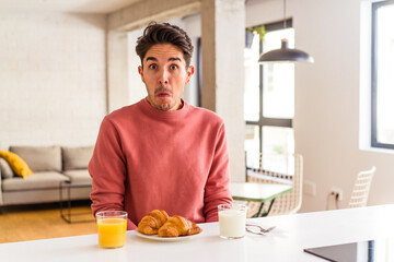 Fototapeta na wymiar Young mixed race man having breakfast in a kitchen on the morning shrugs shoulders and open eyes confused.