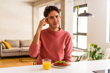 Fototapeta na wymiar Young mixed race man having breakfast in his kitchen pointing temple with finger, thinking, focused on a task.