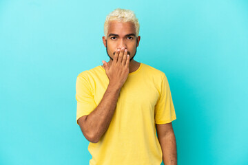 Young Colombian handsome man isolated on blue background covering mouth with hand