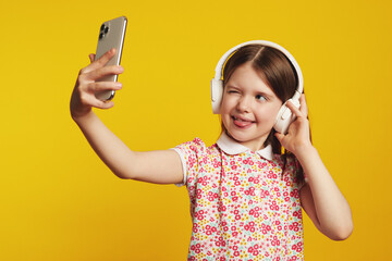 Little girl kid shows tongue and blinks eye while takes selfie on smartphone device, listening music in headphones, stands against yellow background