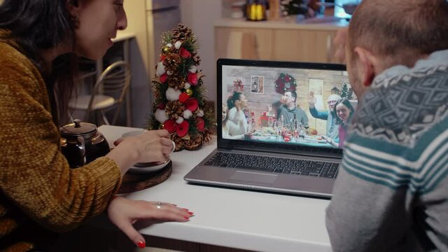 Festive couple talking to family on video call at christmas dinner. Man and woman using laptop for remote conference online, enjoying holiday celebration on internet. People speaking to relatives