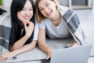Pajamas party. Happy teenage girls. Home movie. Digital lifestyle. Pretty smiling asian caucasian young women laying floor covering plaid with laptop light room interior.