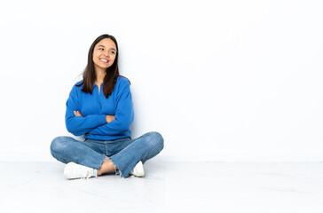 Young mixed race woman sitting on the floor isolated on white background with arms crossed and happy