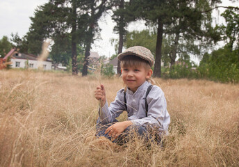 a boy in vintage clothes sits in a field with dry grass, a rural landscape