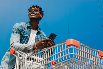 Afro black man with a shopping cart and a phone in his hands, makes purchases online, background...