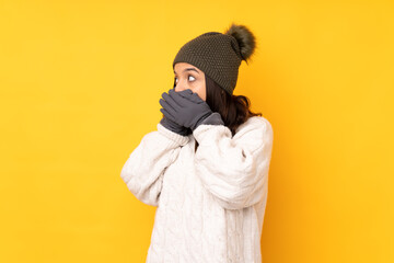 Young woman with winter hat over isolated yellow background covering mouth and looking to the side