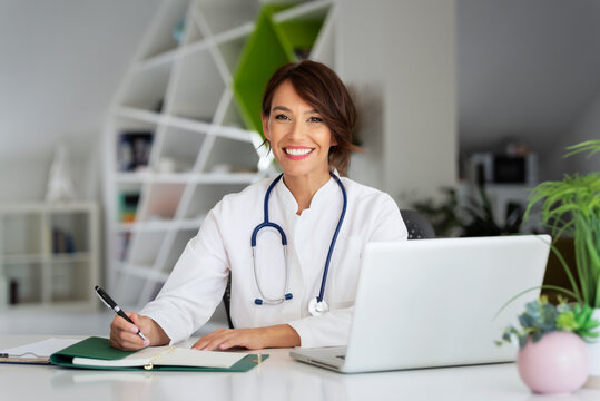 Smiling Female Doctor Using Laptop And Writing Something While Sitting At Doctor's Office