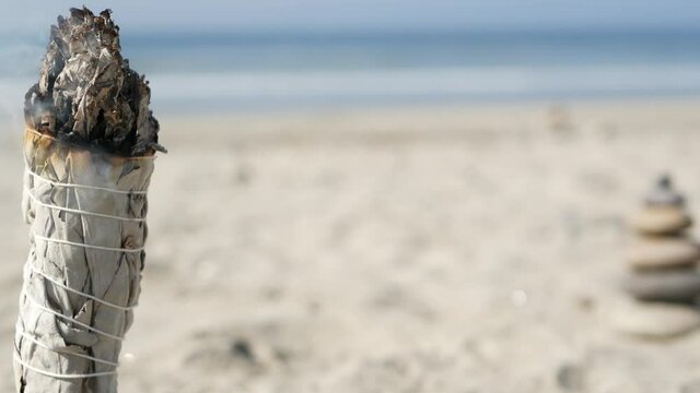 Dried white sage smoke, dry smudge stick burning, soft focus bokeh. Aroma smudging close up on ocean beach, aromatherapy meditation by sea water waves on sandy coast. Yoga, peace, harmony and ayurveda