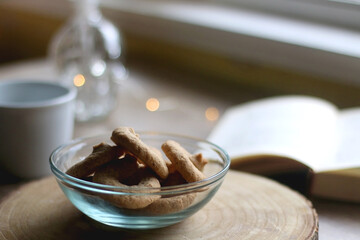 Bowl of cookies, cup of hot beverage, open book and lit candles on a table. Selective focus.