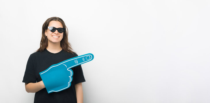 Young woman is wearing sunglasses, foam fan glove and pointing aside over white background.