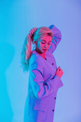 Stylish beautiful young girl in fashionable clothes with a bright coat in the studio on a multicolored neon pink light