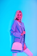 Happy glamorous young smiling woman in fashionable bright business outfit look with blue blazer and handbag posing in studio on colored neon pink light background
