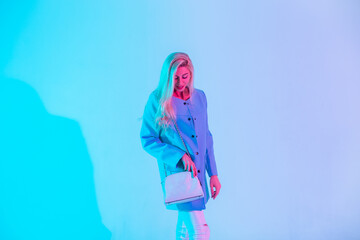 Happy young blonde girl in a fashionable blue elegant blazer with a white stylish leather handbag stands in the studio on neon light with pink